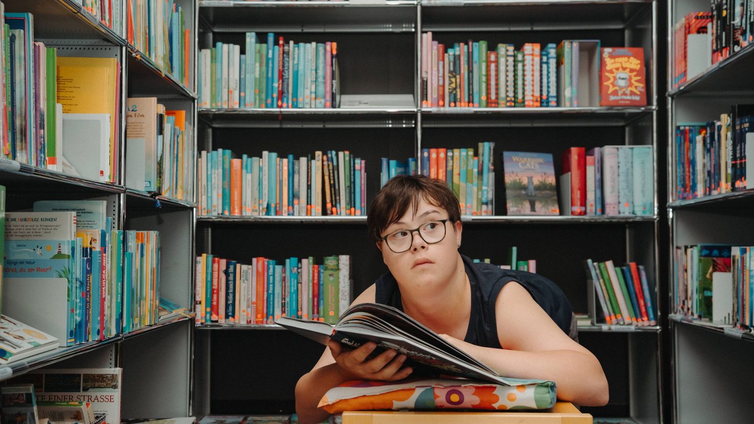 Mädchen mit Unterstützungsbedarf liegt umgeben von Bücherregalen und liest in einem Buch - Teilhabe und Bildung in der Stiftung MBF