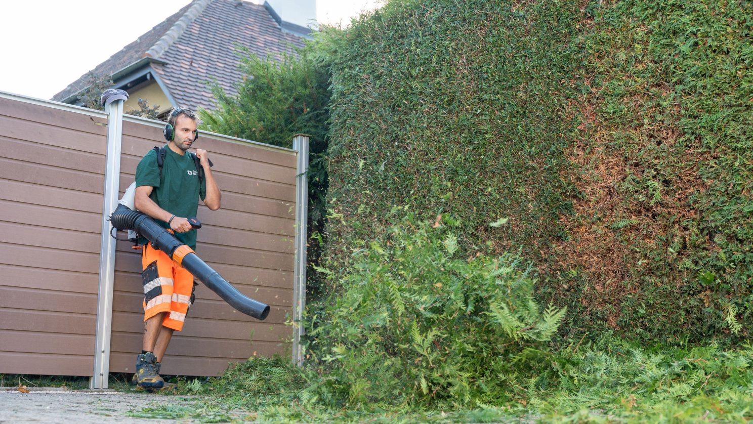 Mitarbeiter der Stiftung MBF entfernt mit einem Laubbläser abgeschnittene Äste und Blätter entlang einer Hecke.