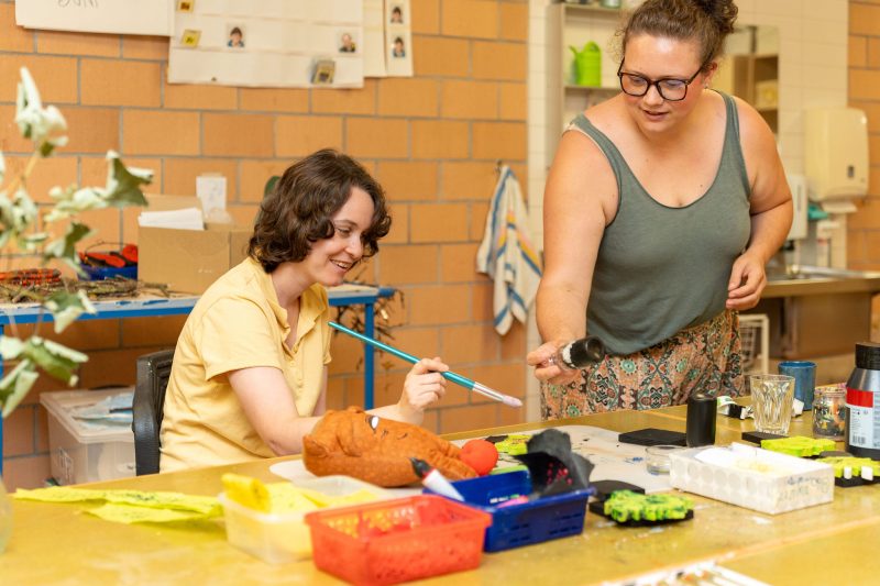 Eine Person sitzt am Tisch und bemalt eine Tonfigur, daneben unterstützt eine Betreuungsperson bei den Arbeitsschritten.