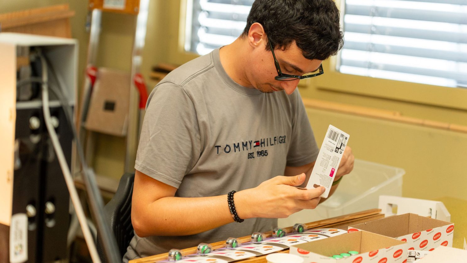 Ein junger Mann legt konzentriert einzelne Artikel in eine Kartonverpackung ein, die vor ihm bereitsteht.