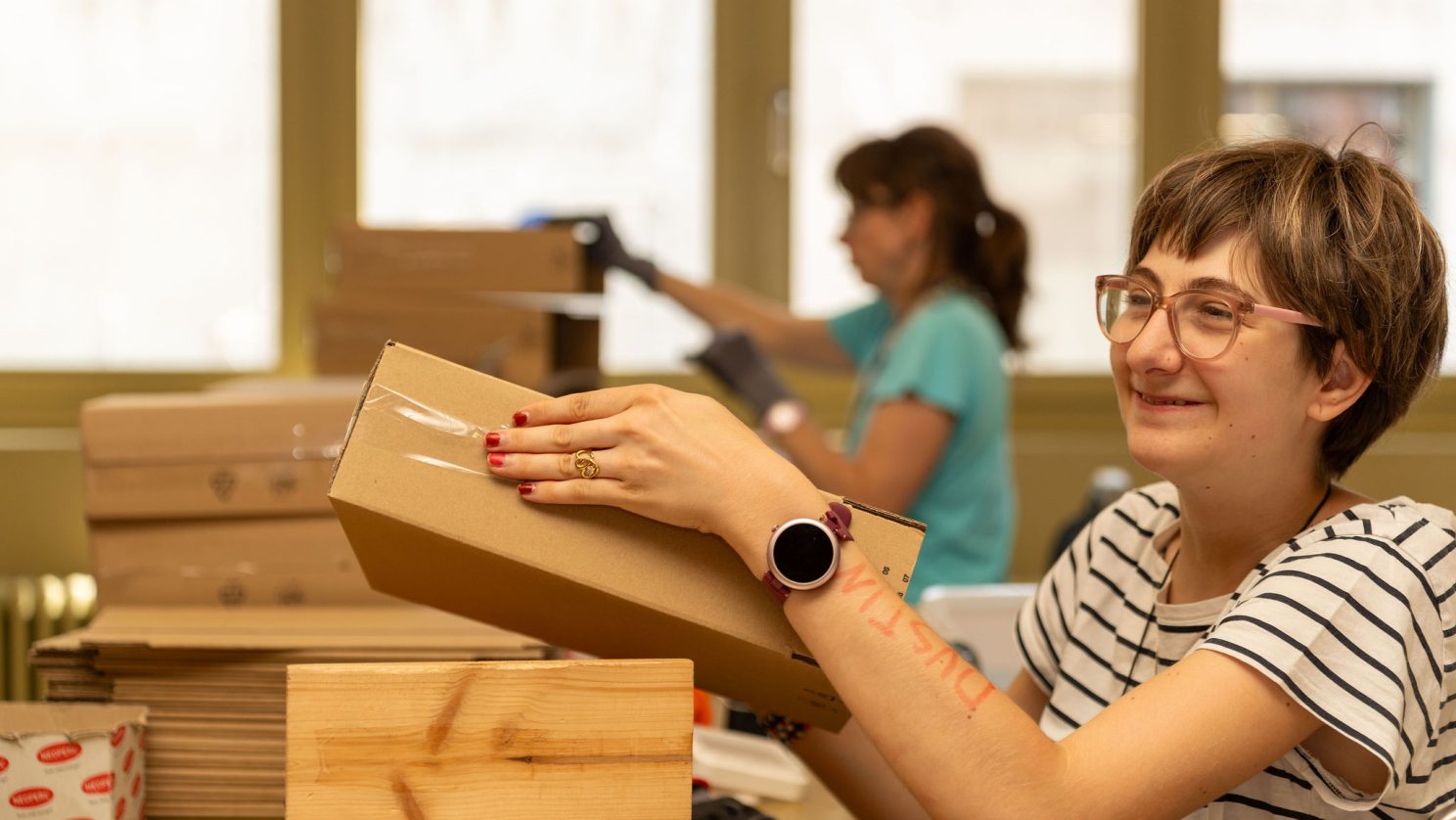 Lächelnde Mitarbeiterin mit Unterstützungsbedarf verpackt einen Karton; hinter ihr arbeitet eine Kollegin am selben Tisch.