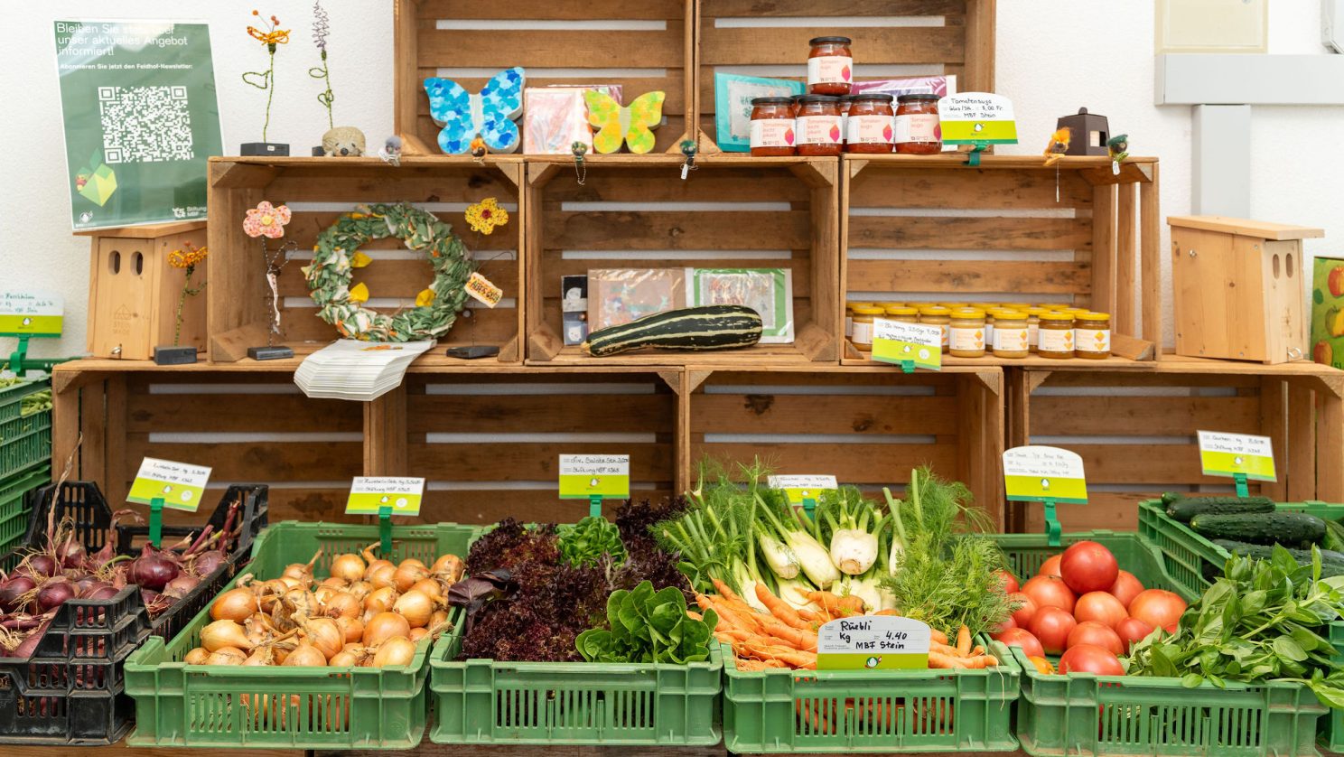 Ein Verkaufsstand im Laden zeigt saisonales Gemüse in grünen Kisten sowie selbstgemachte Produkte und Deko in offenen Holzboxen im Hintergrund.