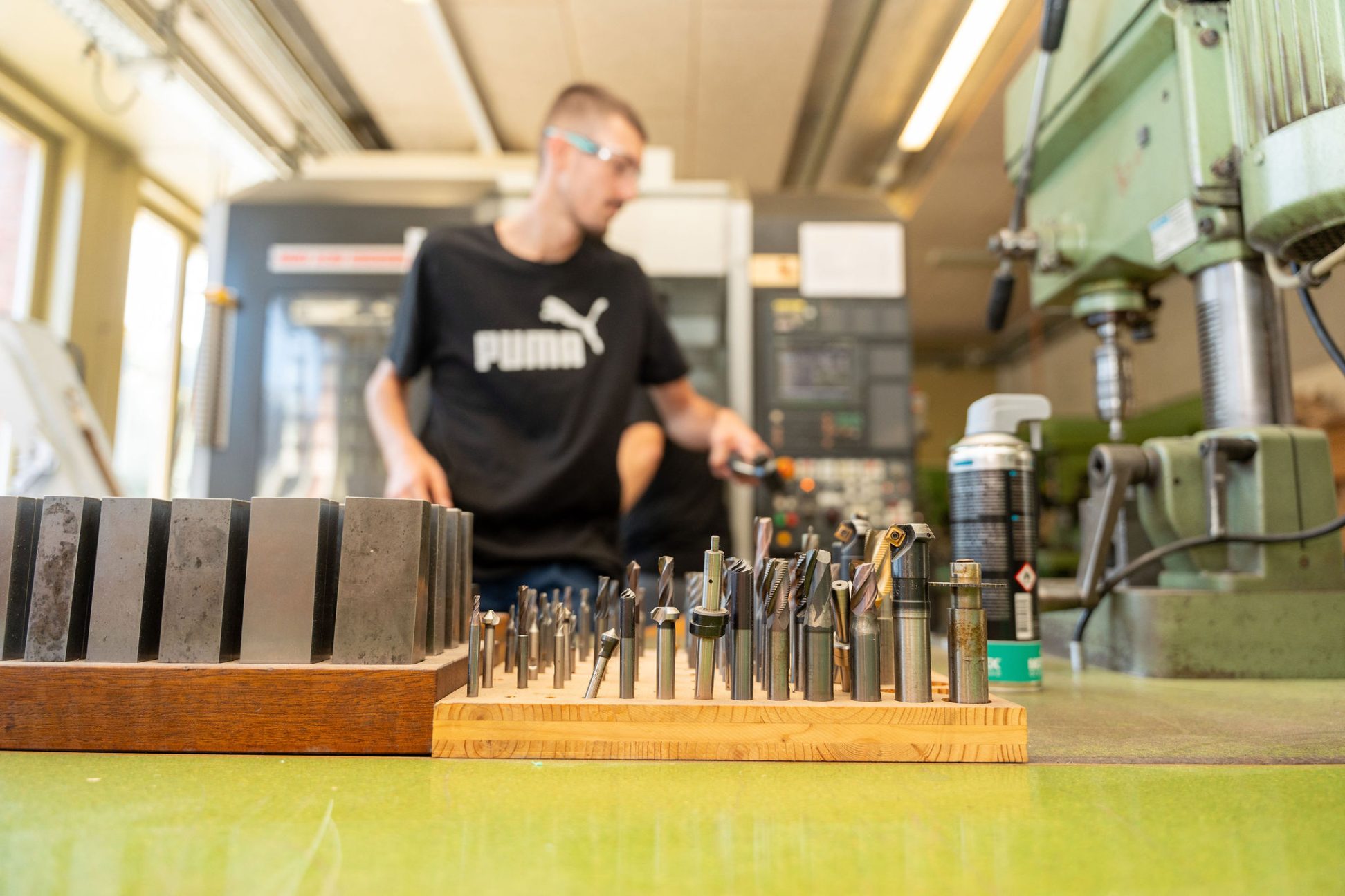 Werkzeugaufsätze und Metallfräser stehen sortiert auf einem Holzblock in einer Werkstatt; im Hintergrund arbeitet eine Person an einer Maschine.