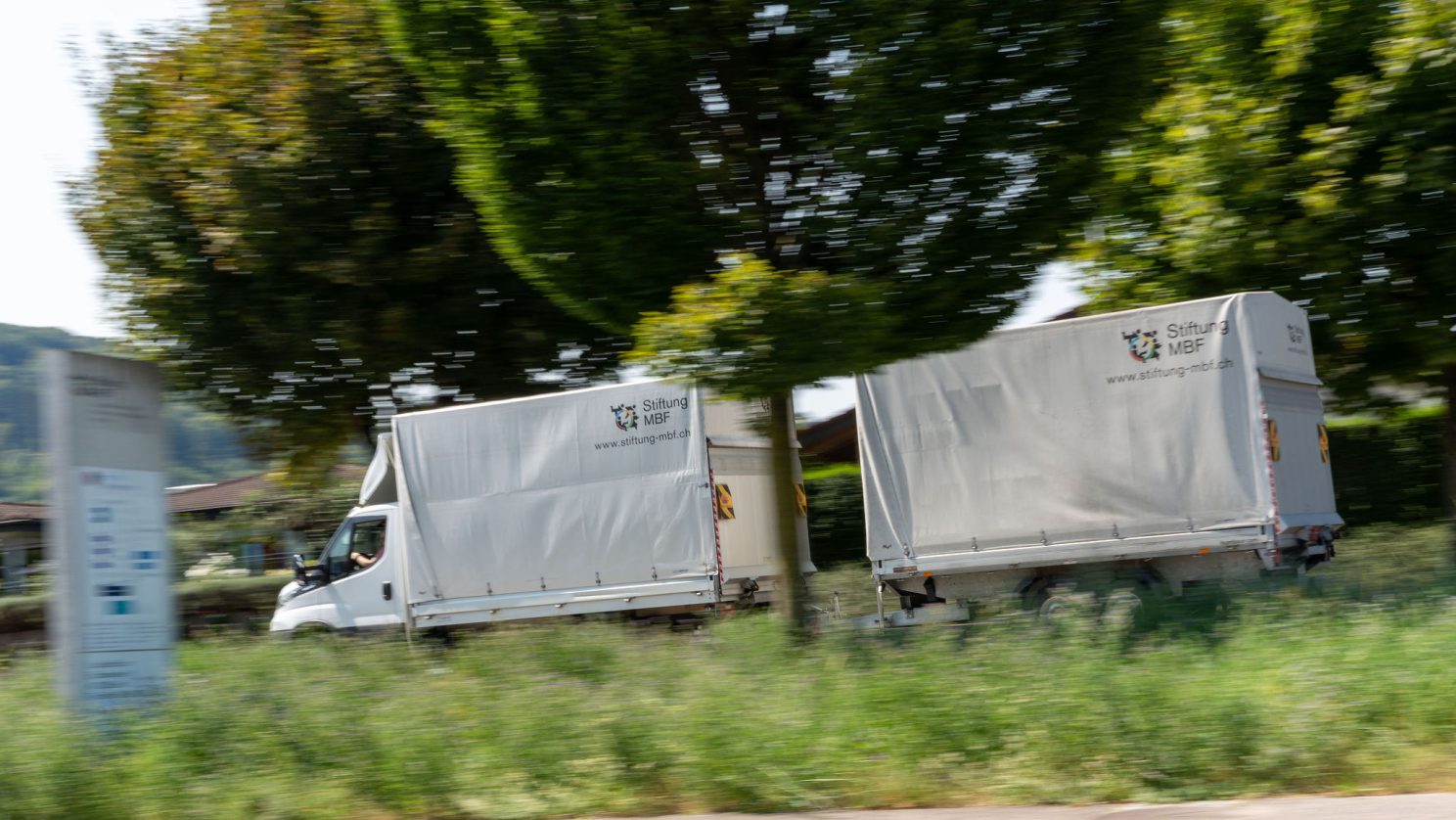 MBF-Transportfahrzeug mit Planenaufbau passiert eine bepflanzte Strassenkante.