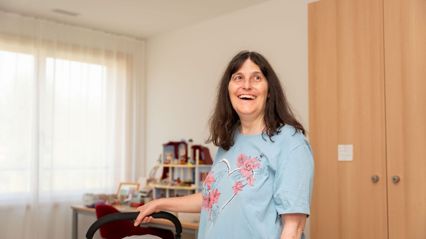 Frau mit Unterstützungsbedarf blickt fröhlich in die Richtung der Kamera, im Hintergrund ihr persönlich eingerichtetes Zimmer.