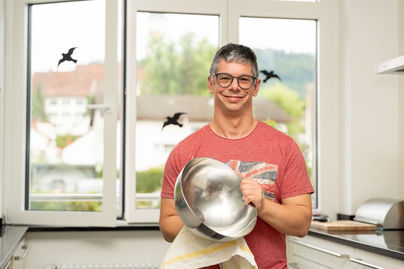 Ein Mann in rotem T-Shirt trocknet in einer hellen Küche eine Metallschüssel und schaut dabei freundlich in die Kamera.