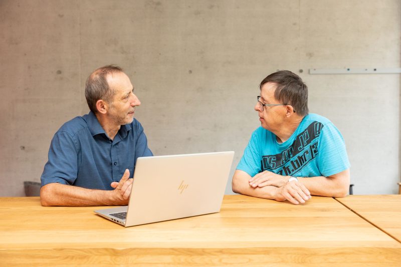 Ein Angestellter der Stiftung MBF spricht mit einem Mitarbeitenden mit Unterstützungsbedarf an einem Tisch über Inhalte am Laptop.