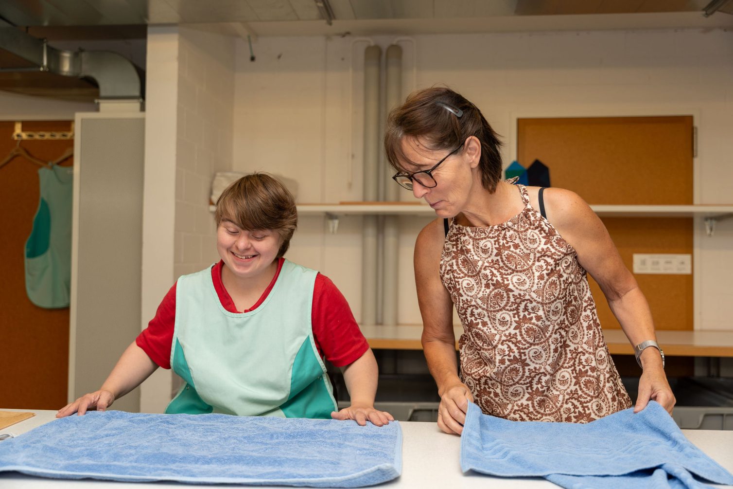 Zwei Frauen legen Handtücher auf einem Tisch zusammen. Die linke jüngere Frau ist eine Person mit Trisonomie 21 und die Dame rechts ist eine Betreuungsperson, die sich unterstützenden mit der jüngeren Dame unterhält.