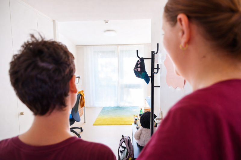 Im Vordergrund sind zwei Frauen, eine jünger, eine älter, von hinten. Sie blicken gemeinsam in einen hellen freundliche Wohnraum