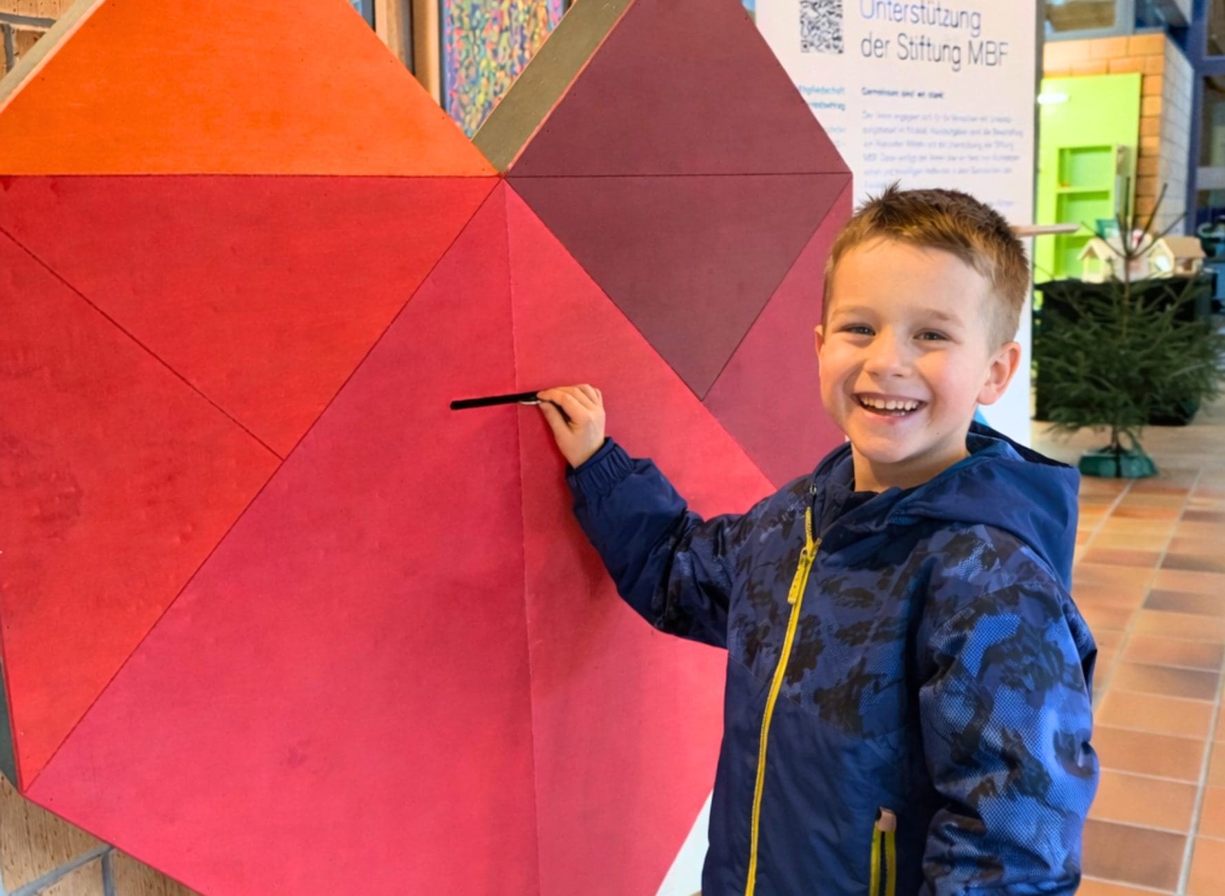 Ein dunkelblonder Junge im Kindergartenalter wirft lächelnd ein Geldstück in ein grosses Spendenherz mit Schlitz aus Holz.