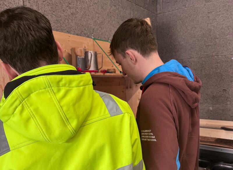 Zwei junge Männer arbeiten an Seite an einer Werkvorrichtung. der Mann rechts ist ein Zivildienstleistende, links ein Mitarbeiter mit Unterstützungsbedarf.